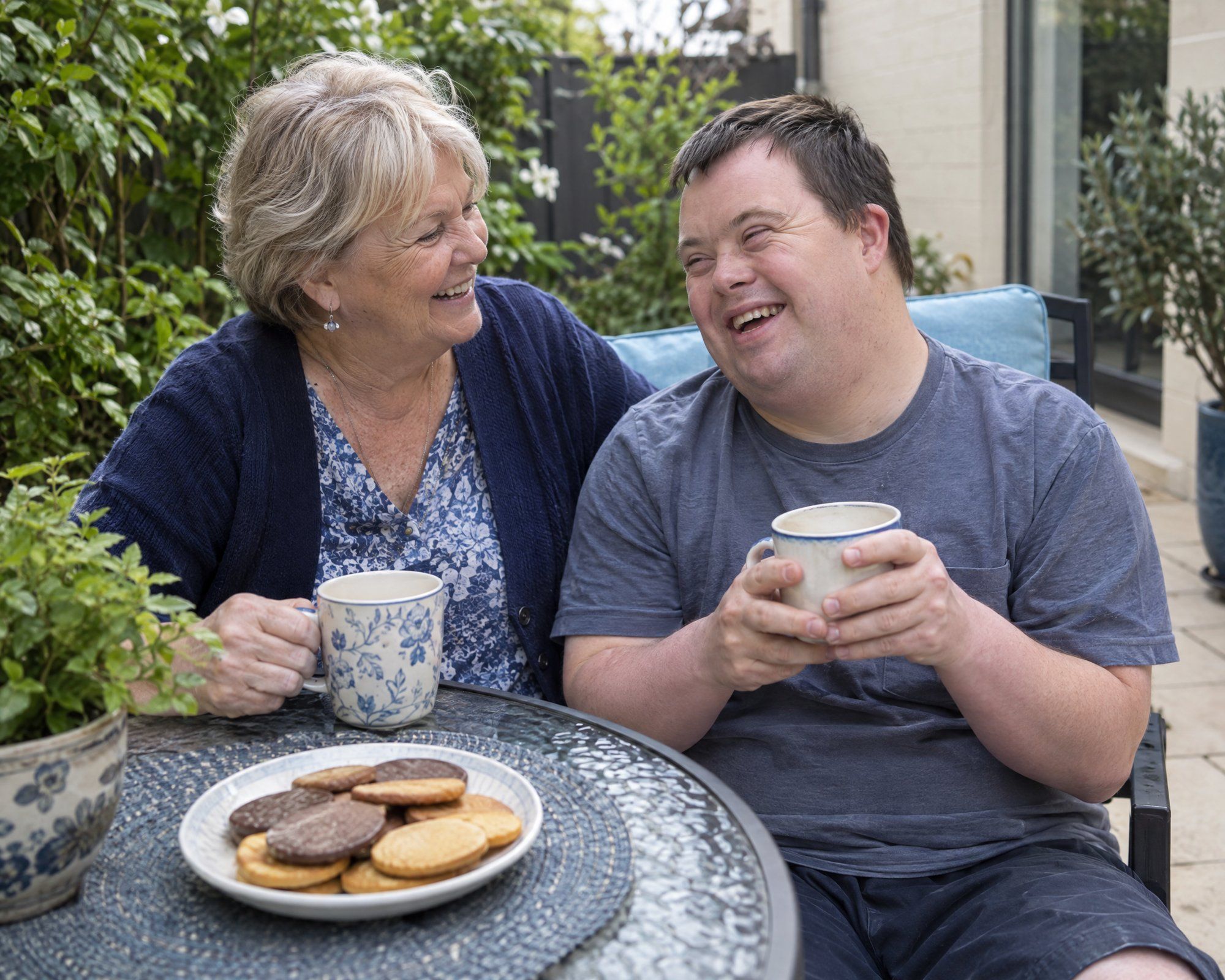 A mother and her adult son share a laugh over coffee in his sunny courtyard